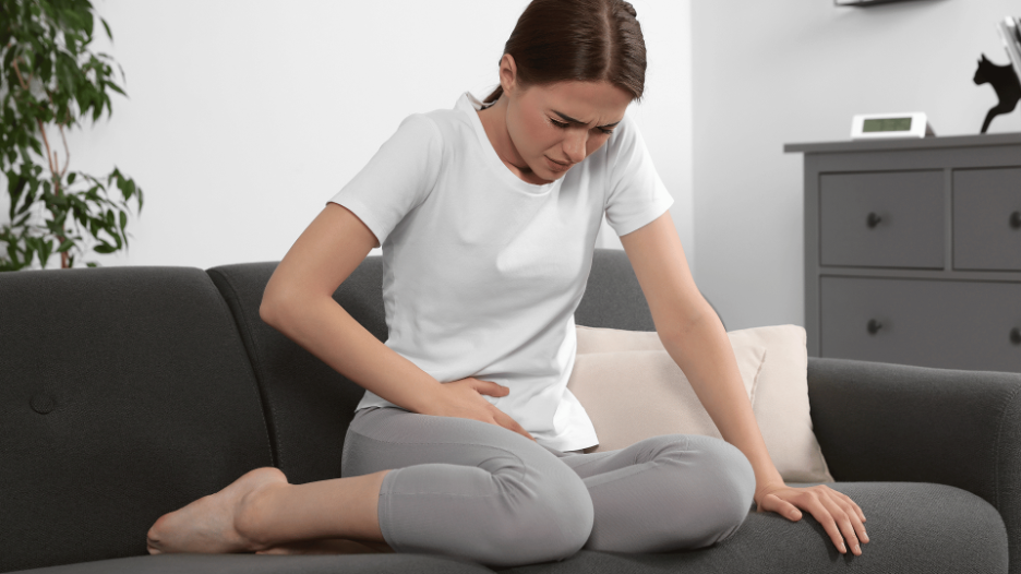 Woman sitting on a couch, holding her lower abdomen in pain, indicating symptoms of a UTI or yeast infection