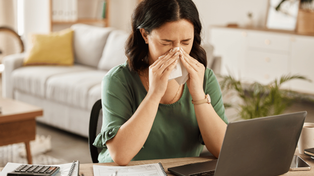 A woman sneezing into a tissue while sitting at her desk is experiencing sinus infection symptoms highlighting the challenges of bacterial vs. viral sinus infections.