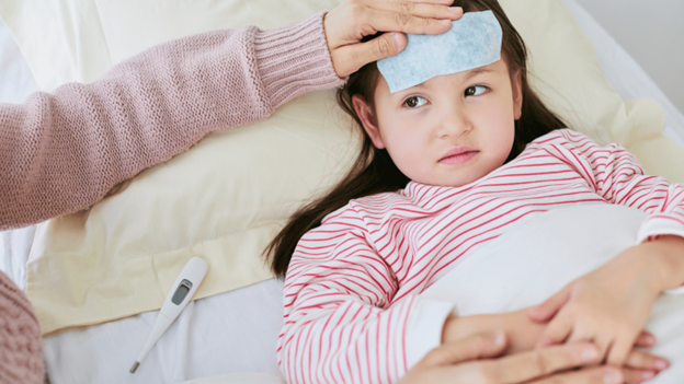 Sick child lying in bed with a fever patch on her forehead and a thermometer nearby, showing symptoms of the flu in children